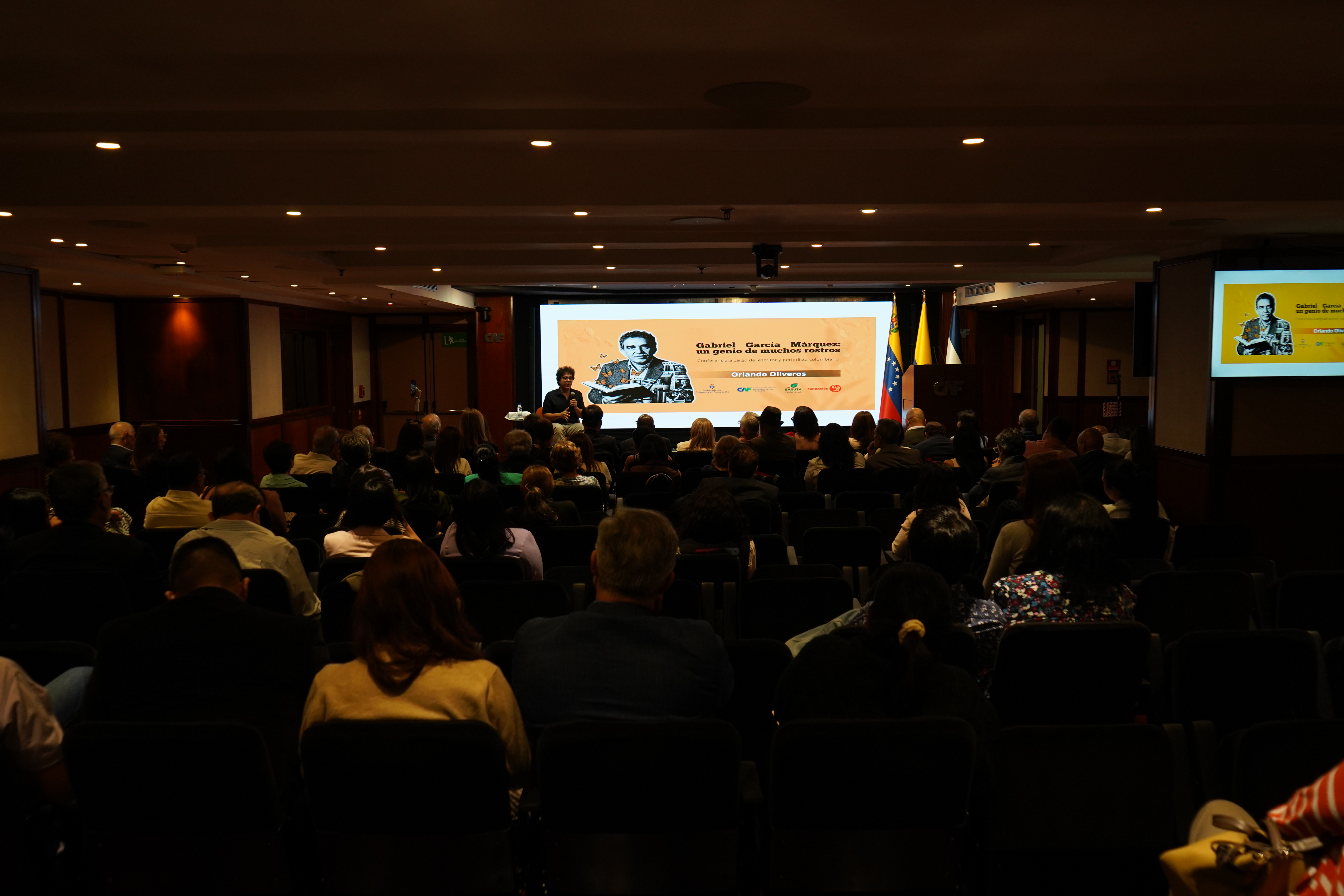 El escritor y periodista cultural colombiano Orlando Oliveros durante su conferencia en el auditorio de CAF, acompañado por el público asistente.